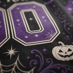 Close-up of a purple and black fabric with a large number 00, white stars, spiderwebs, swirls, and a jack-o'-lantern in a Halloween pattern.