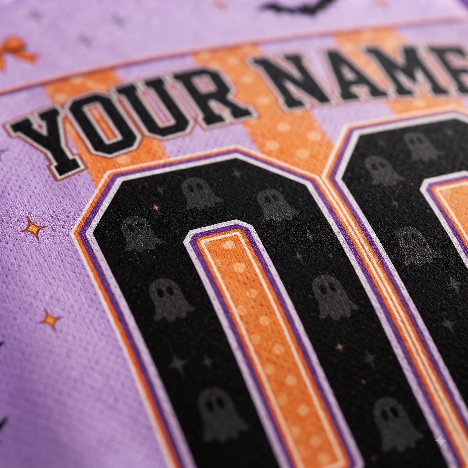 Close-up of a purple athletic jersey with a Halloween-themed graphic featuring the text 'YOUR NAME', black numbers outlined in orange and purple, and repeating ghost motifs.