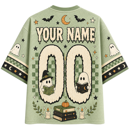 A sage green short-sleeve t-shirt with a Halloween-themed graphic design on the back, featuring 'YOUR NAME 00' for personalization, cartoon ghosts reading books, bats, stars, and a crescent moon.