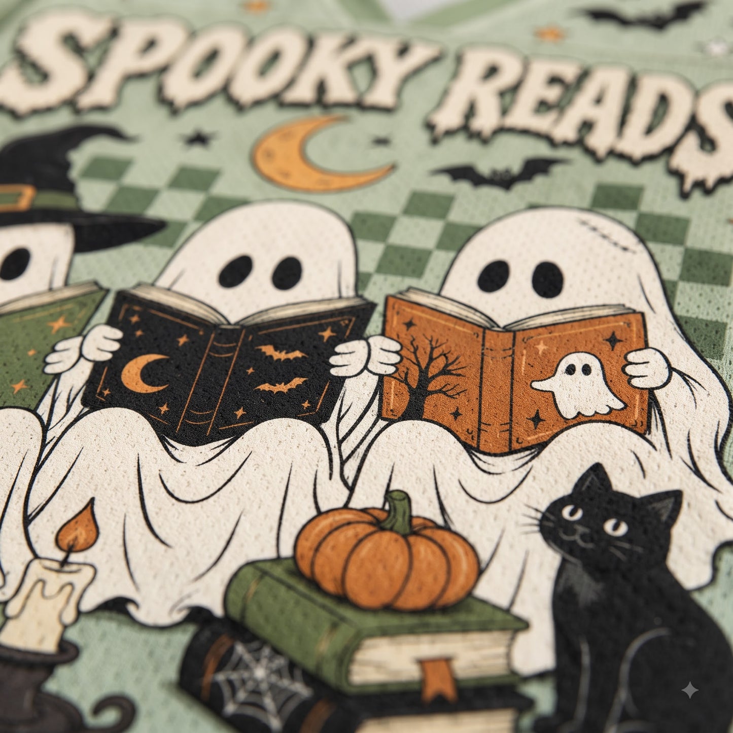 A close-up of a Halloween-themed book cover or printed design featuring two ghosts reading books, a black cat, stacked books, a candle, pumpkins, bats, and the words “SPOOKY READS” on a green patterned background.