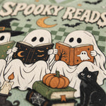 A close-up of a Halloween-themed book cover or printed design featuring two ghosts reading books, a black cat, stacked books, a candle, pumpkins, bats, and the words “SPOOKY READS” on a green patterned background.