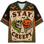 A short-sleeve V-neck jersey-style shirt with a Halloween graphic print. The design features the words 'STAY CREEPY' in dripping black letters, a green hand holding a skull, a skeleton hand holding a carved orange pumpkin, bats, stars, and leopard-print border details on a cream background.