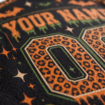Close-up of a custom embroidered design on a black fabric, featuring an orange leopard print number 0 with green and white outlines, orange stars, and orange text reading 'YOUR NAM'.