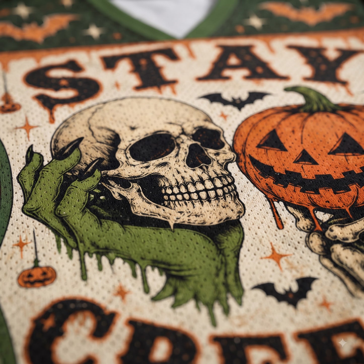 Close-up of a Halloween-themed printed garment featuring a skull, green skeletal hand, orange jack-o'-lantern, bats, stars, and distressed text on a cream mesh fabric.