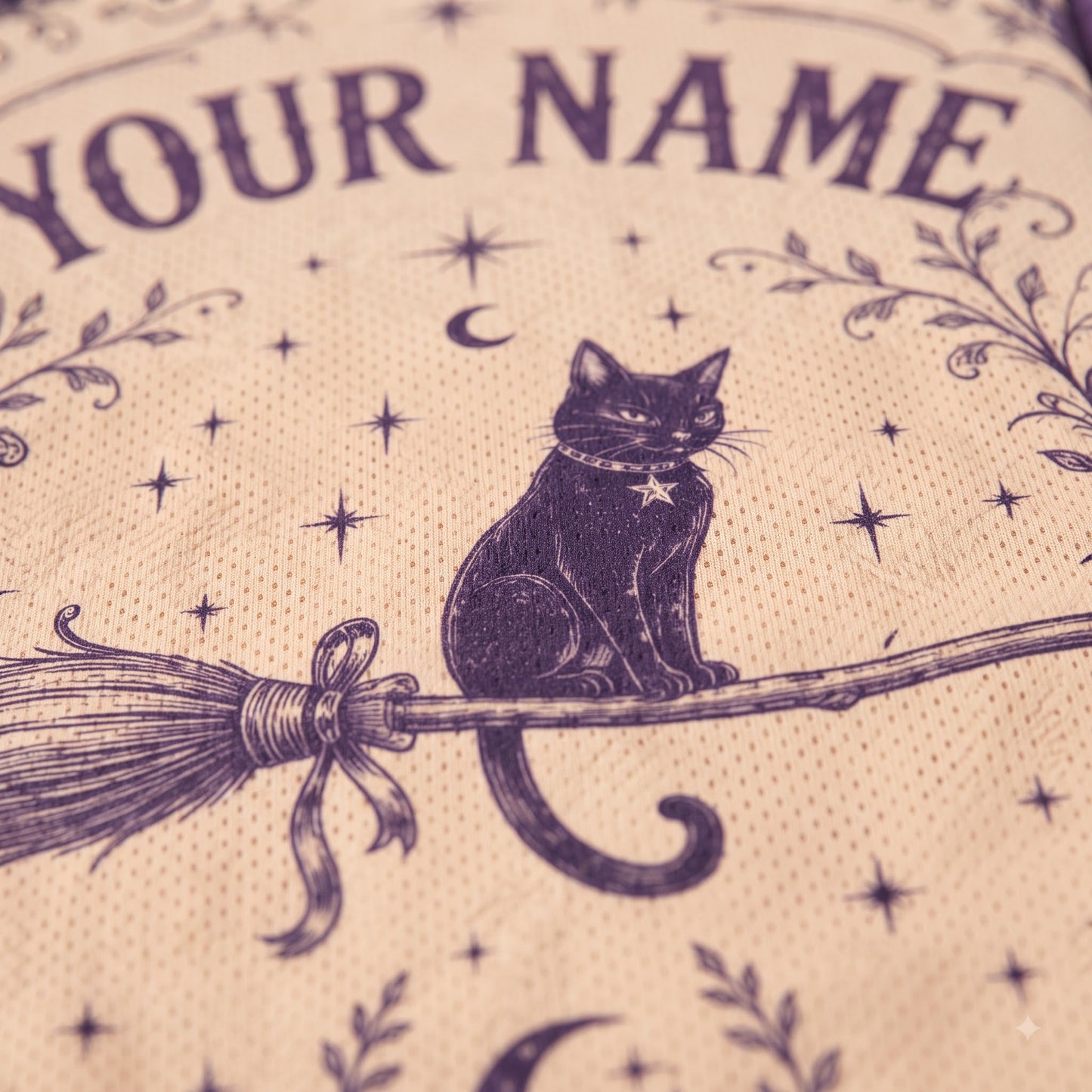 Close-up of a beige fabric item printed with a dark purple witch-themed design, including a black cat on a broomstick, stars, crescent moon, floral motifs, and the text 'YOUR NAME'.