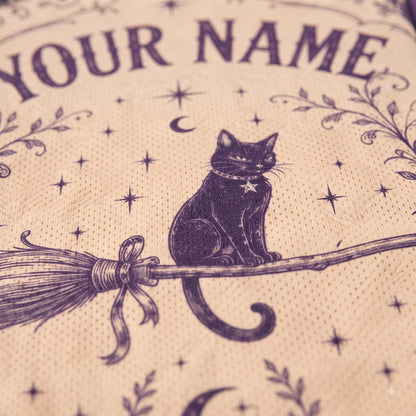 Close-up of a beige fabric item printed with a dark purple witch-themed design, including a black cat on a broomstick, stars, crescent moon, floral motifs, and the text 'YOUR NAME'.