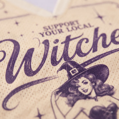 A close-up of a beige woven fabric surface printed with dark purple text reading 'SUPPORT YOUR LOCAL Witch' and an illustrated witch wearing a pointed hat.