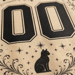 Close-up of a beige printed surface with large black '00' numerals, a black cat silhouette, star motifs, and decorative flourishes.