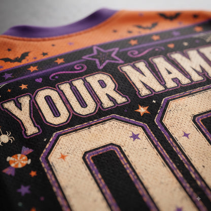 Close-up of a Halloween-themed custom jersey back with 'YOUR NAME' in cream lettering outlined in purple on a black background, decorated with orange stars, purple bats and stars, and a purple collar.