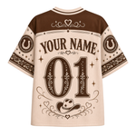A short-sleeve western-style jersey shirt shown from the back, featuring a cream base with dark brown panels and graphics. The design includes 'YOUR NAME' and '01' with decorative floral and star motifs, horseshoes on the sleeves, and a cowboy hat near the hem.