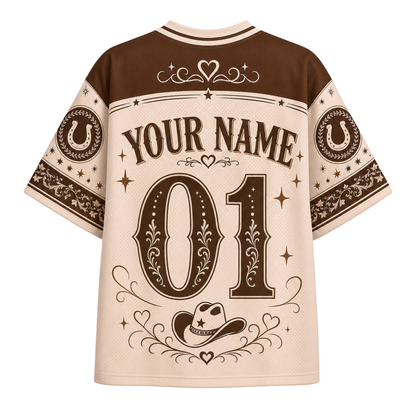 A short-sleeve western-style jersey shirt shown from the back, featuring a cream base with dark brown panels and graphics. The design includes 'YOUR NAME' and '01' with decorative floral and star motifs, horseshoes on the sleeves, and a cowboy hat near the hem.