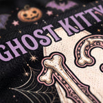 Close-up of a black knit garment featuring a Halloween-themed graphic with the words 'GHOST KITTY' in lavender, a cream-colored skeleton letter 'K', pink and cream bones, spiderwebs, stars, and a pumpkin motif.