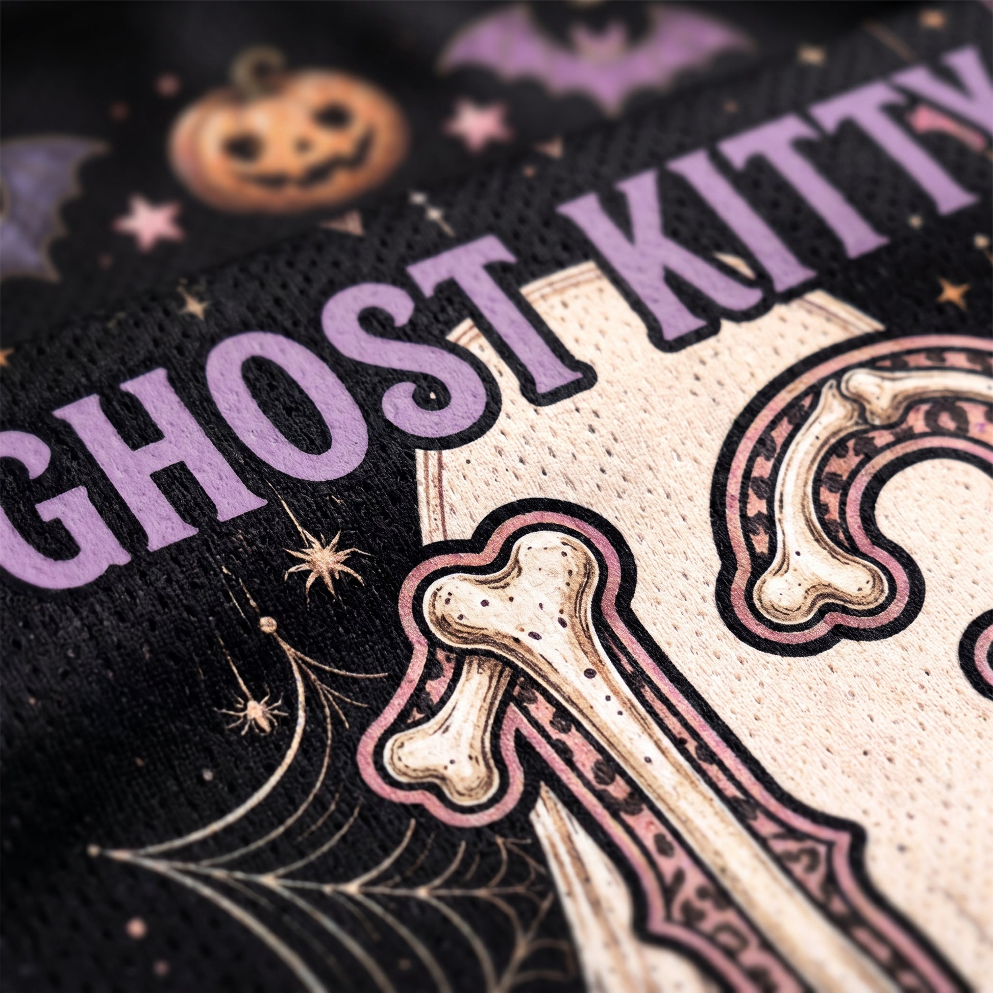 Close-up of a black knit garment featuring a Halloween-themed graphic with the words 'GHOST KITTY' in lavender, a cream-colored skeleton letter 'K', pink and cream bones, spiderwebs, stars, and a pumpkin motif.