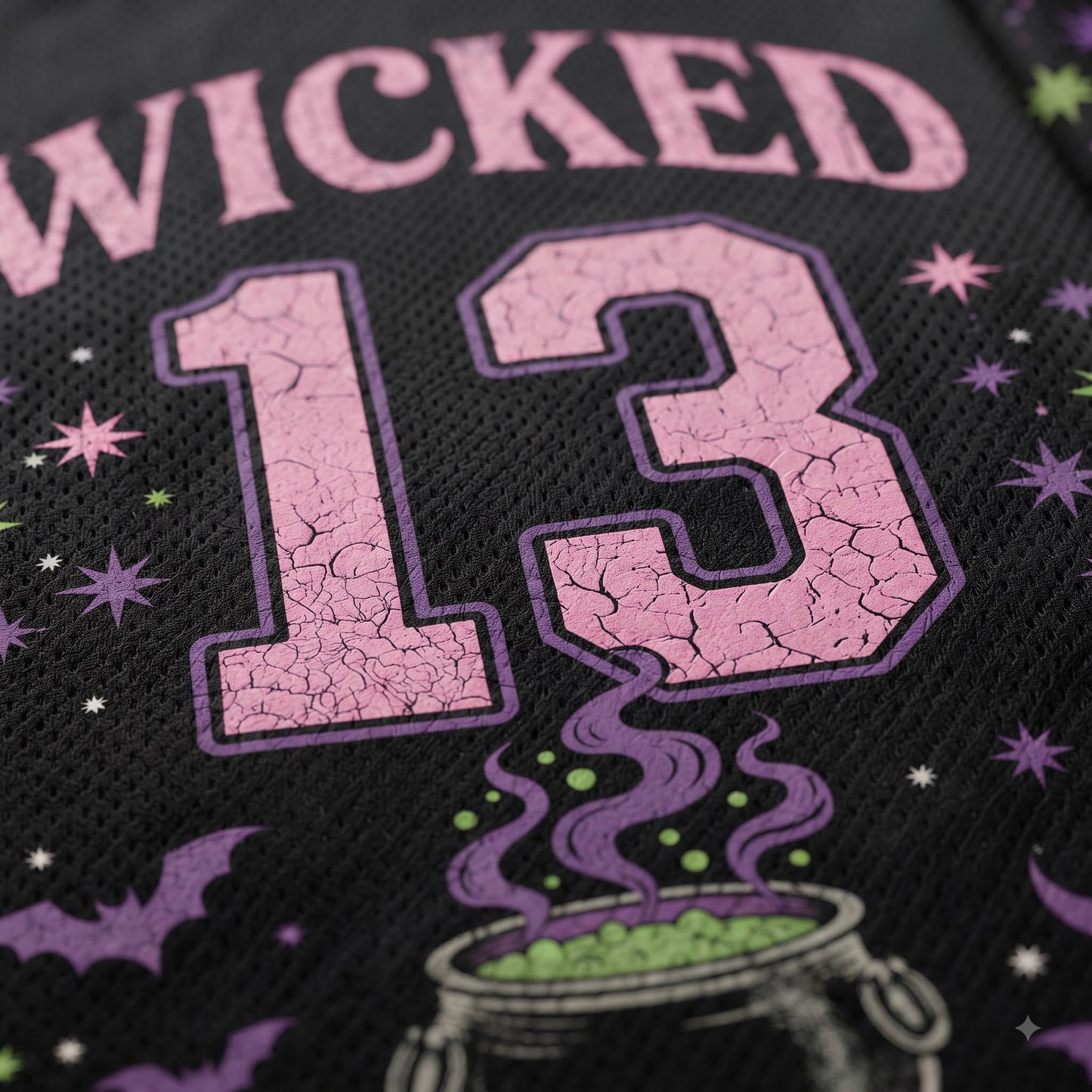 Close-up of a black mesh fabric featuring a pink and purple cracked-texture graphic with the number 13, the word "WICKED" above it, and small witch-themed illustrations including stars, bats, and a cauldron with green potion.