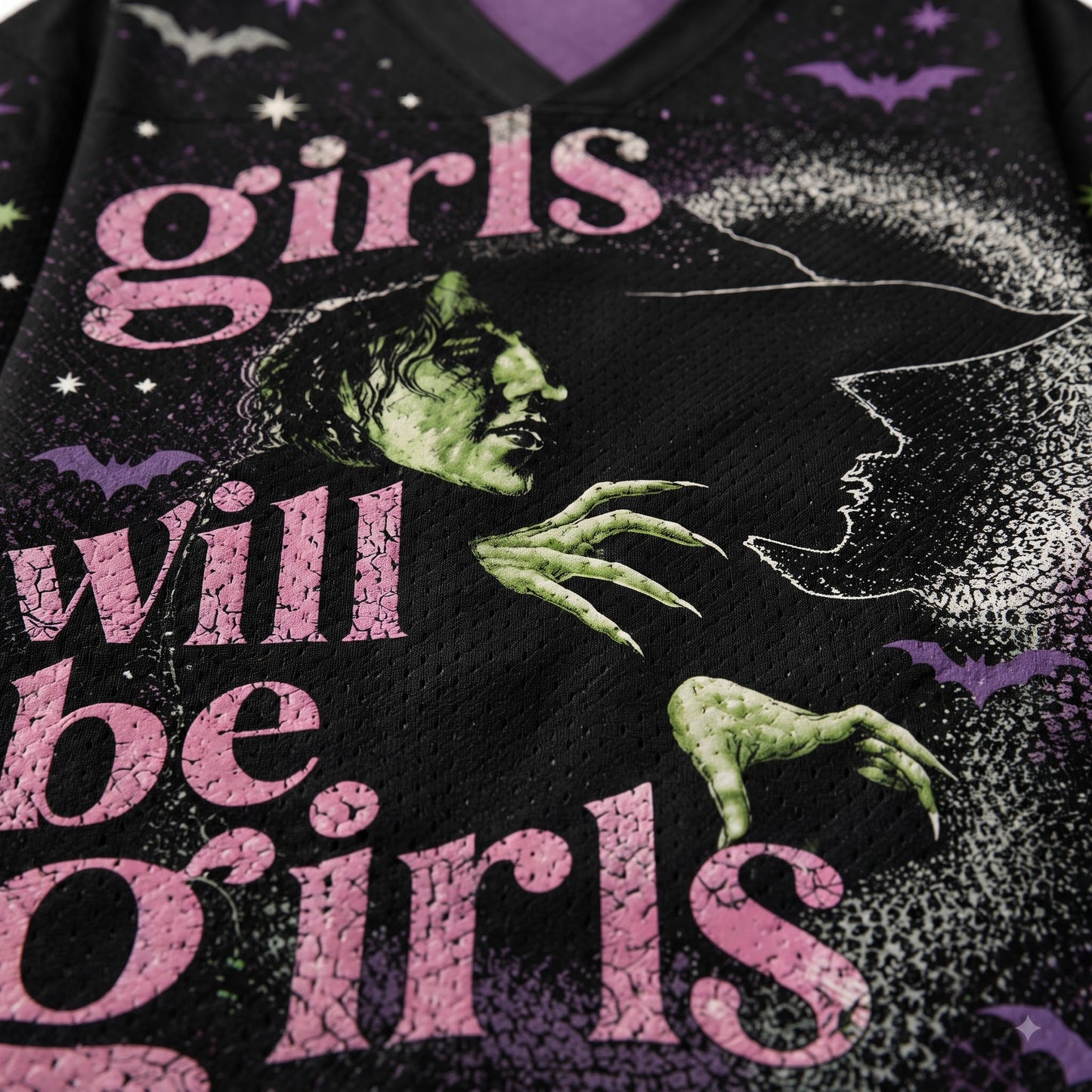 Close-up of a black mesh graphic shirt featuring the phrase 'girls will be girls' in pink distressed lettering, with a green witch face, green clawed hand, white bat silhouettes, and purple stars.