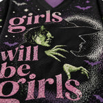 Close-up of a black mesh graphic shirt featuring the phrase 'girls will be girls' in pink distressed lettering, with a green witch face, green clawed hand, white bat silhouettes, and purple stars.