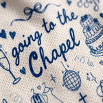 Close-up of a textured cream fabric printed with navy blue script reading 'going to the Chapel' and small wedding-themed illustrations including hearts, a cake, a champagne glass, and a disco ball.
