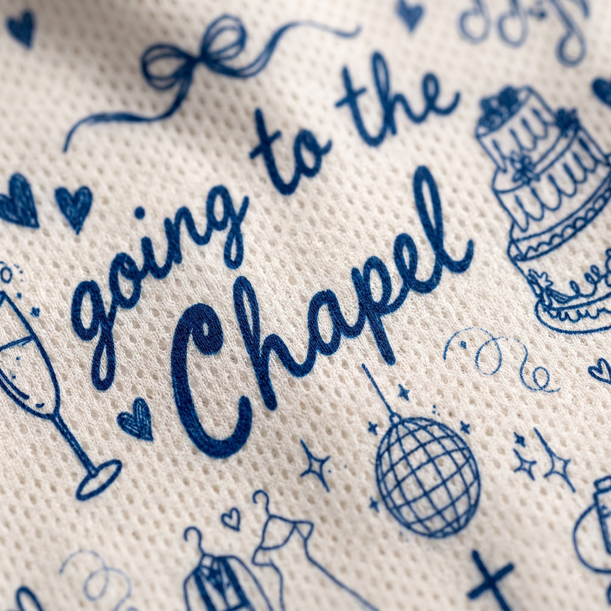 Close-up of a textured cream fabric printed with navy blue script reading 'going to the Chapel' and small wedding-themed illustrations including hearts, a cake, a champagne glass, and a disco ball.