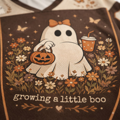 Close-up of a brown graphic print on a garment featuring a white ghost holding a jack-o'-lantern and a drink, surrounded by flowers and butterflies, with the text 'growing a little boo'.