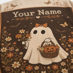 Close-up of a personalized Halloween-themed fabric item, likely a bag or pouch, featuring a cream-colored ghost graphic with the text 'Your Name' at the top. The design includes a pumpkin basket, flowers, butterflies, and bats on a dark brown background.