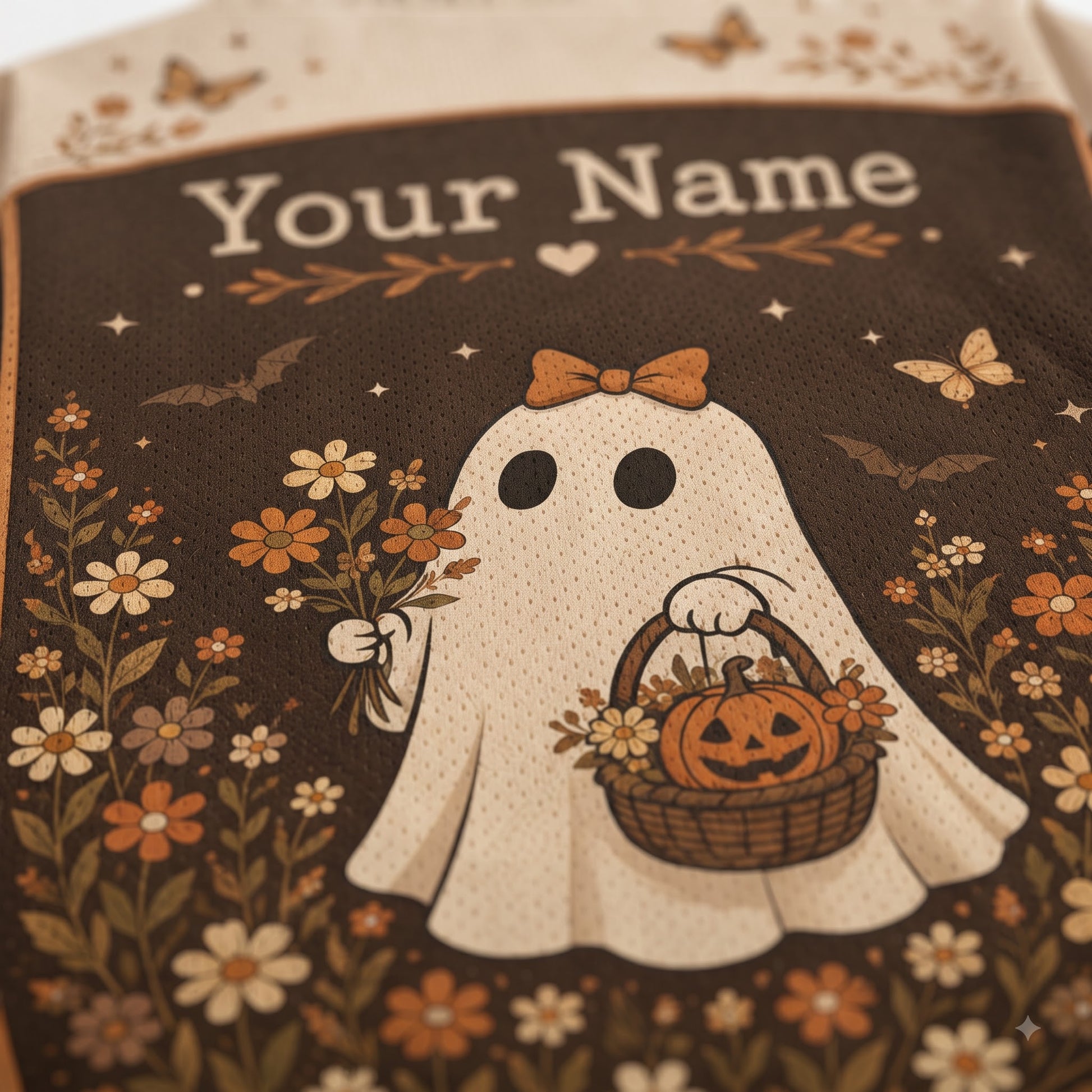 Close-up of a personalized Halloween-themed fabric item, likely a bag or pouch, featuring a cream-colored ghost graphic with the text 'Your Name' at the top. The design includes a pumpkin basket, flowers, butterflies, and bats on a dark brown background.