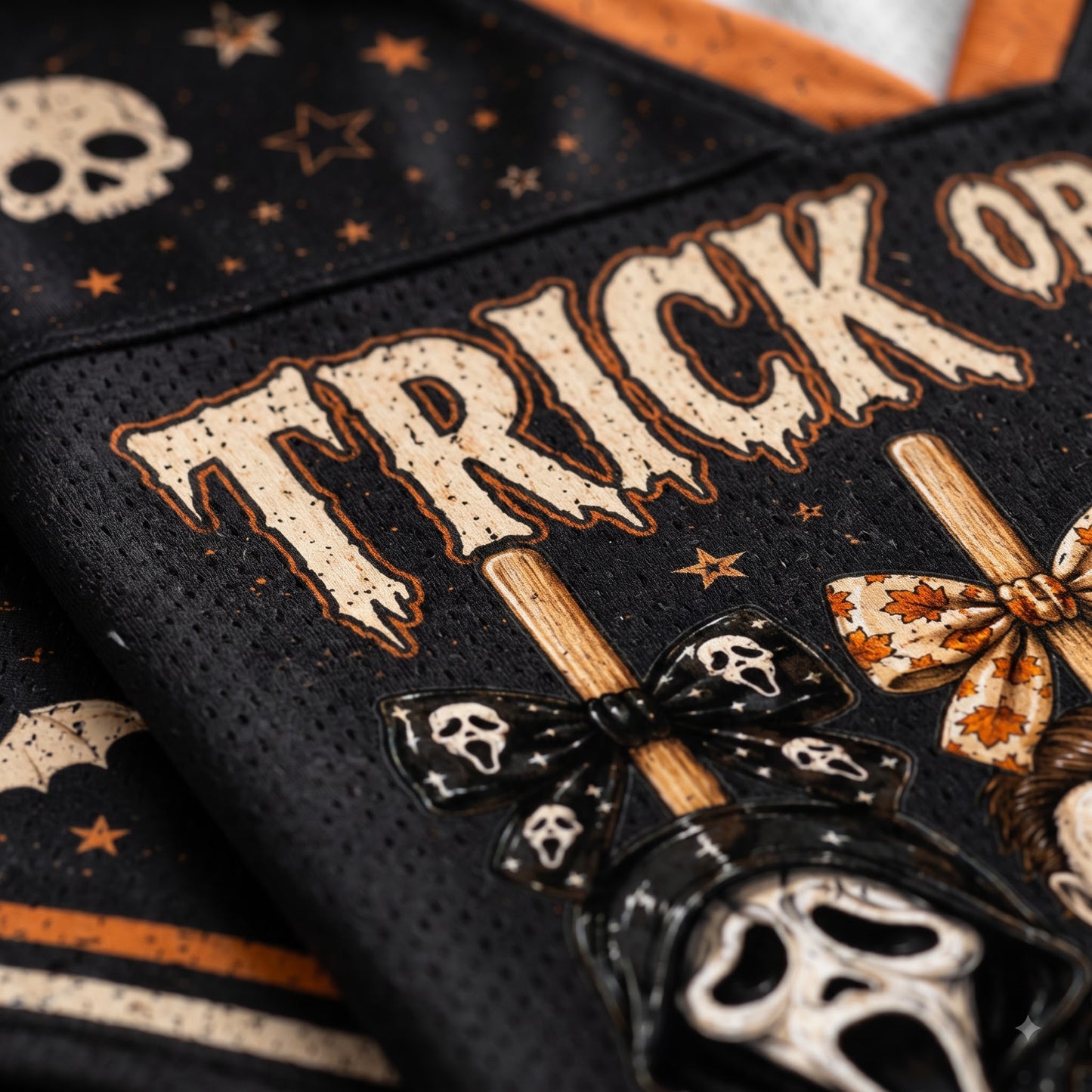 Close-up of a black mesh fabric with Halloween-themed graphics, including the words “TRICK OR TREAT,” skulls, stars, and decorative bows in beige, orange, and black.