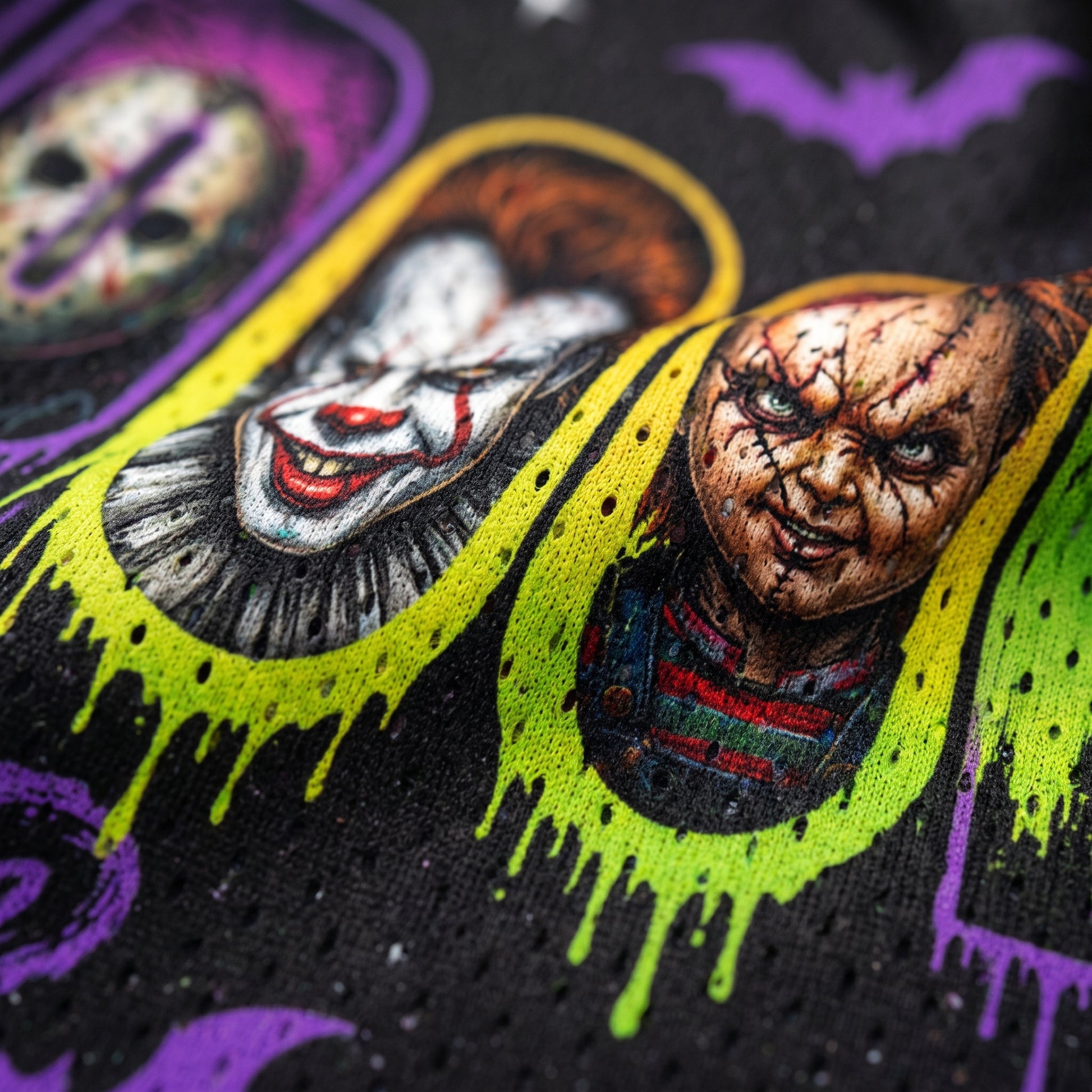 Close-up of a black fabric printed with horror character faces, neon yellow drip outlines, purple bat silhouettes, and splattered details.