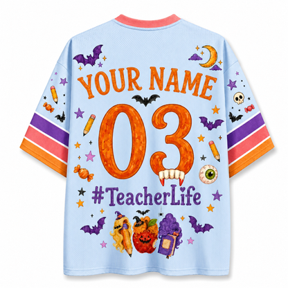 A light blue short-sleeve t-shirt with a colorful Halloween-themed back graphic. The design features the text 'YOUR NAME 03 #TeacherLife' in orange and purple, surrounded by illustrations of bats, pumpkins, candy corn, a witch hat, and stars.