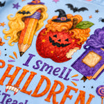 Close-up of a light blue fabric featuring a colorful Halloween-themed graphic with the text 'I smell CHILDREN' in orange and purple, surrounded by illustrated elements including a pencil with curly blonde hair, a jack-o'-lantern apple, a purple potion bottle, bats, and stars.