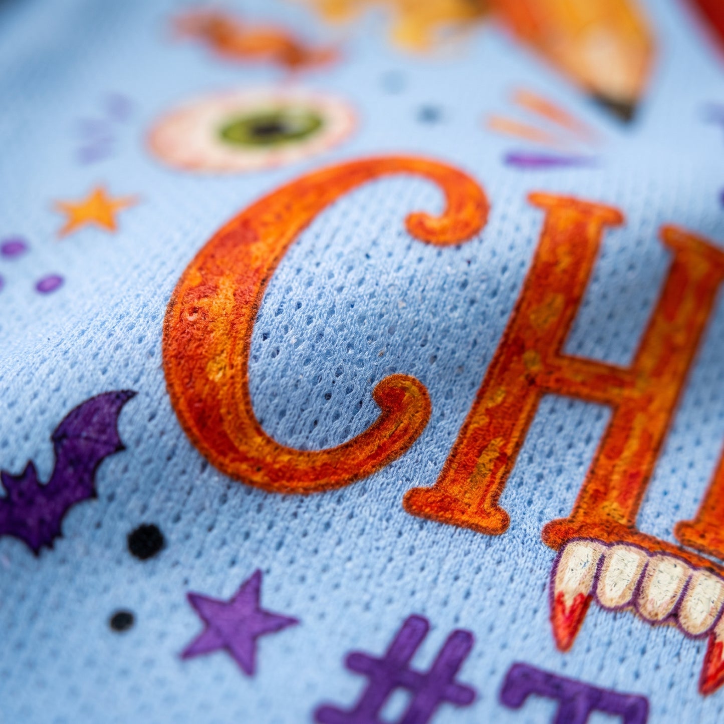 Close-up of a light blue knit fabric with embroidered Halloween-themed lettering and motifs, including an orange 'C', purple stars and bats, and white fangs.