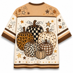 A short-sleeve t-shirt with a large autumn-themed pumpkin graphic on the back. The design features five pumpkins in various patterns (checkered, leopard print, polka dot, plaid) on a cream background, decorated with stars, leaves, and swirls in brown and gold tones.