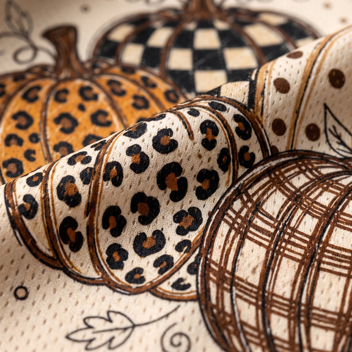 A close-up of a fabric or wallpaper pattern featuring stylized pumpkins in autumn animal and geometric prints, including leopard spots, checkered patterns, polka dots, and plaid lines on a cream background.
