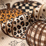 A close-up of a fabric or wallpaper pattern featuring stylized pumpkins in autumn animal and geometric prints, including leopard spots, checkered patterns, polka dots, and plaid lines on a cream background.