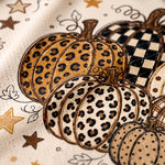 Close-up of a pumpkin print pattern on a textured fabric, featuring illustrated pumpkins with leopard print, checkered pattern, and polka dots in beige, brown, black, and white.