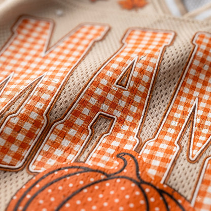 Close-up of a fabric product featuring large orange and white gingham check letters outlined in brown, with an orange pumpkin graphic at the bottom on a beige mesh background.