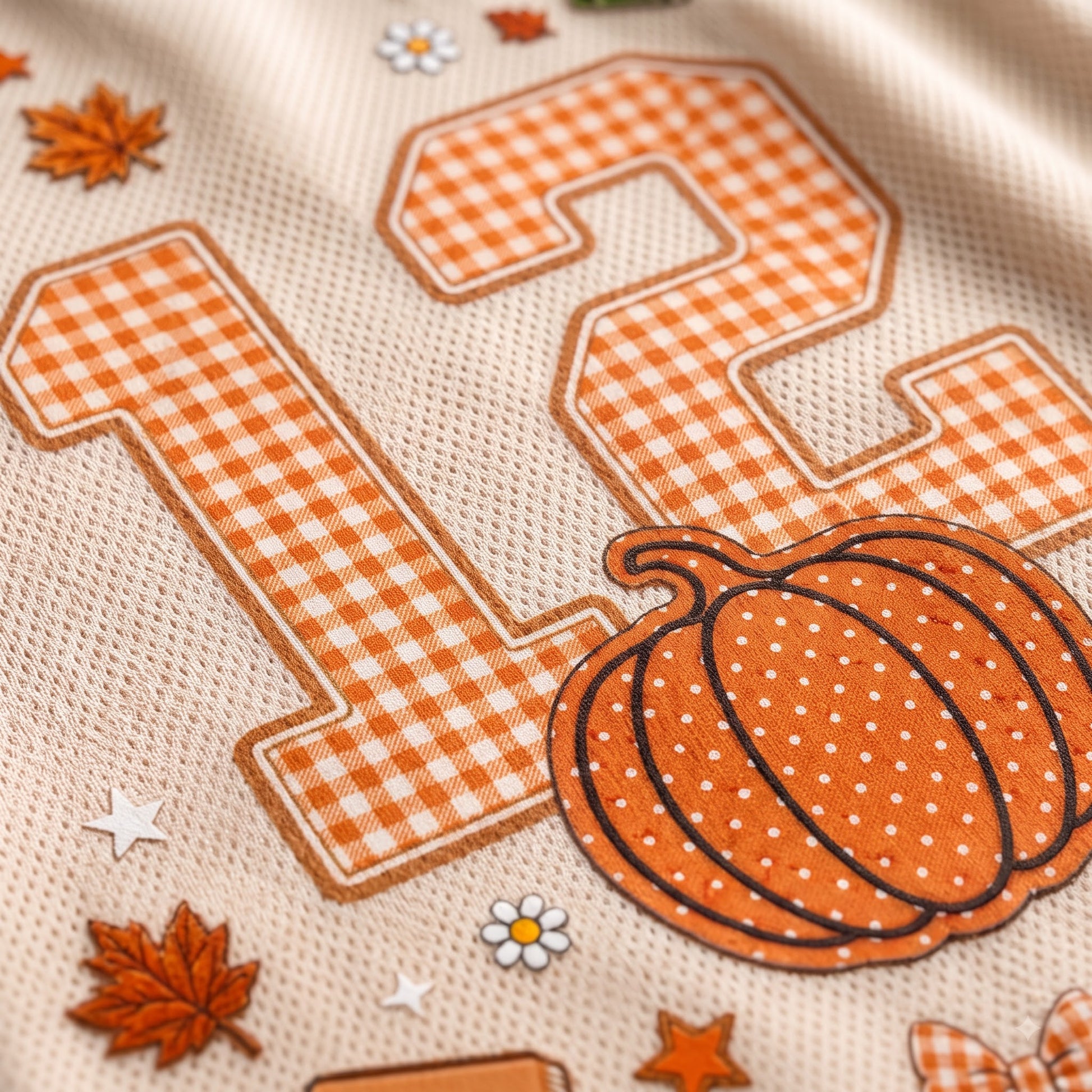 Close-up of a beige textured fabric with a large orange-and-white gingham number 12, an orange pumpkin patch with white dots, small orange leaves, stars, and white dais flowers.
