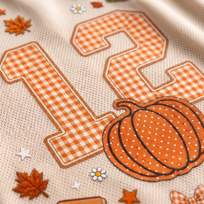 Close-up of a beige textured fabric with a large orange-and-white gingham number 12, an orange pumpkin patch with white dots, small orange leaves, stars, and white dais flowers.