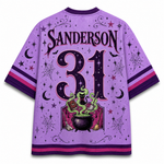 A purple short-sleeve jersey-style shirt shown from the back, featuring the name SANDERSON and the number 31 in large decorative lettering. The design includes a witch illustration with a cauldron, green smoke, stars, moons, bats, and spiderwebs, plus pink and purple striped sleeve and hem accents.