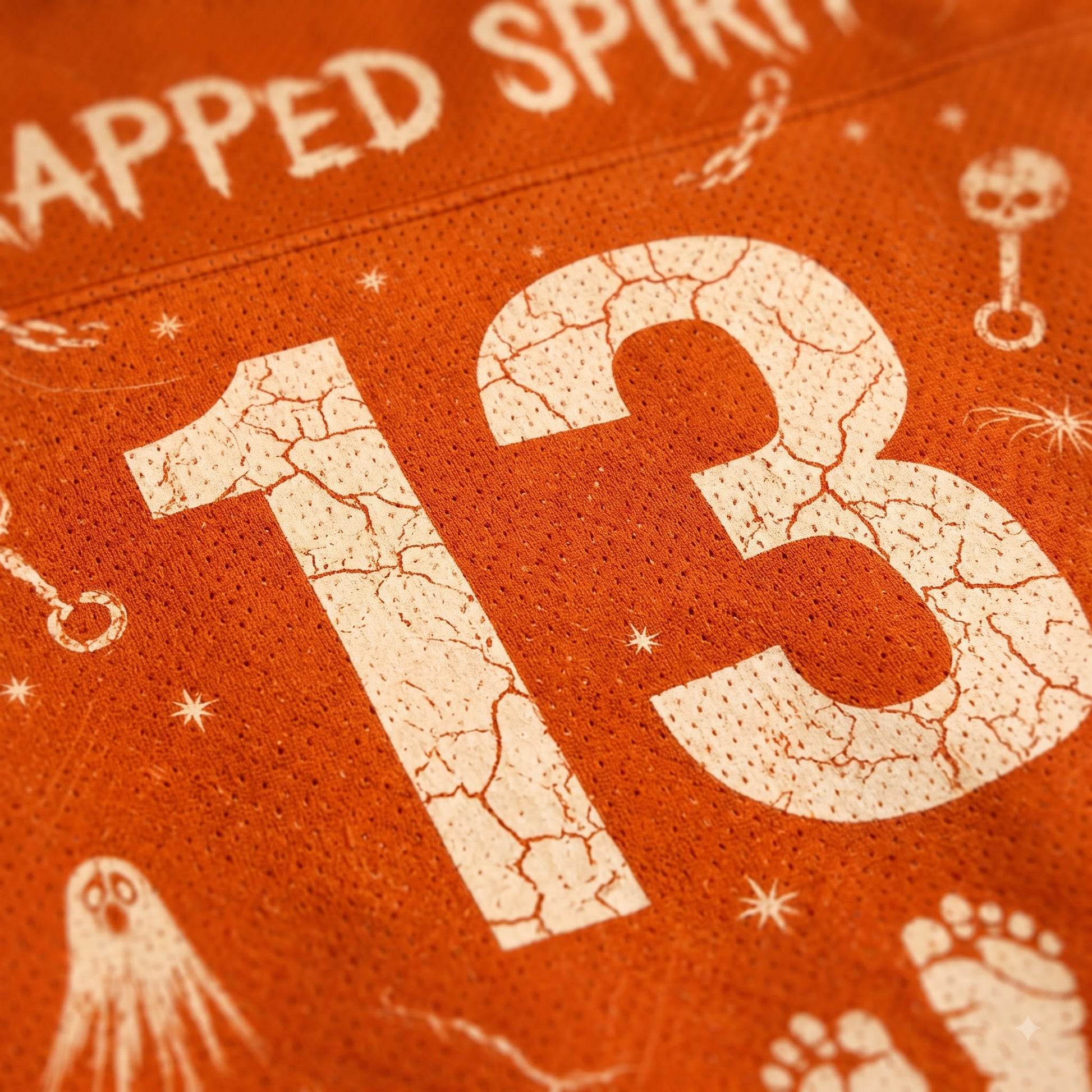 Close up view of distressed cracked number 13, TRAPPED SPIRIT text, ghost, footprint, and key print on rust orange mesh fabric.