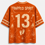An orange short-sleeve t-shirt shown from the back, featuring a Halloween-themed graphic print in a light cream color. The design includes the words 'TRAPPED SPIRIT' across the upper back, the number '13' in the center, and spooky illustrations such as ghosts, keys, stars, webs, and footprints.