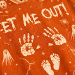 Close-up of orange fabric printed with distressed white Halloween graphics, including the text 'LET ME OUT!', handprints, footprints, ghosts, and stars.